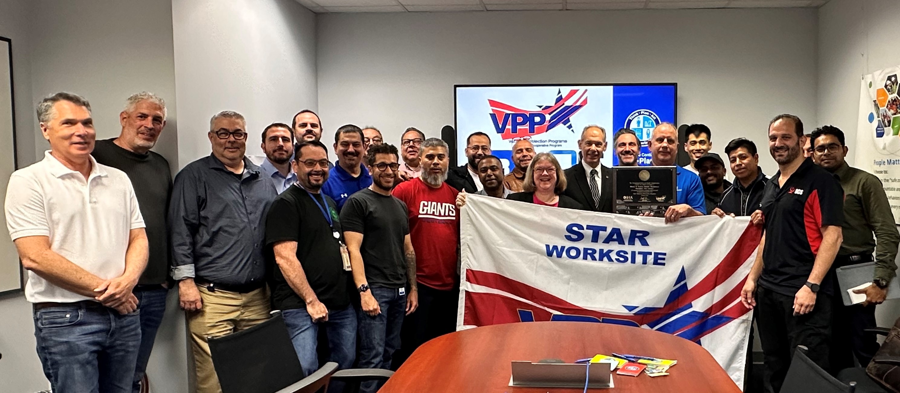 OSHA Regional Administrator, Richard Mendelson with OSHA Area Director, Lisa Levy and Safety Manager Edward Williams hold the VPP Star flag and plaque with more than 20 Black & personnel standing behind them.