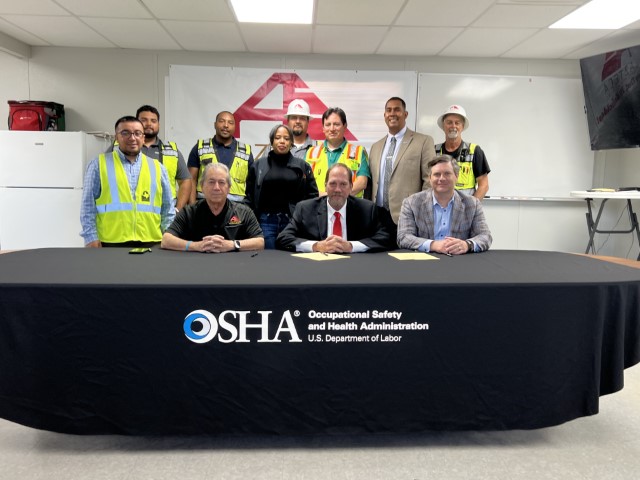 In an office seated at a table, Gregory R. Wynn, OSHA, with Luis Spinola and Jerry McPhail, Azteca Enterprises, signing the agreement. Standing behind them are Eduardo Jimenez, OSHA and seven workers with Aztec Enterprises.