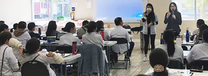 Training room with 2 speakers, one using microphone and the other wearing mask, presenting a slideshow